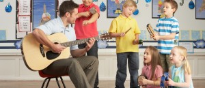 Male Teacher Playing Guitar With Pupils Having Music Lesson In Classroom